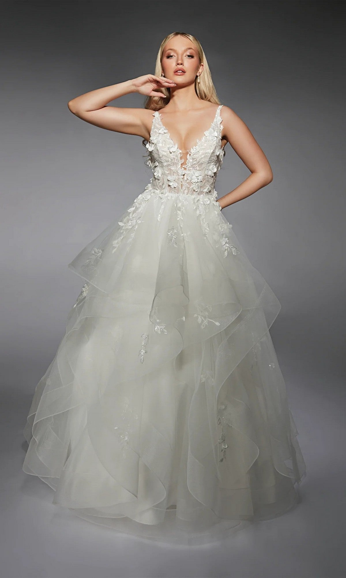 A woman poses against a gray background wearing the Alyce 7131, a long ivory dress with a sleeveless design, deep V-neckline, 3D floral lace appliques, and a layered tulle skirt.